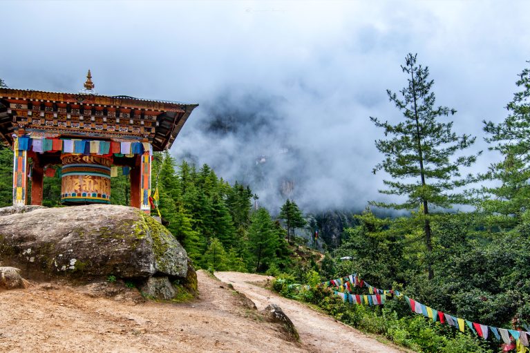 A trail leading towards Bhutanese Monastery lined with prayer flags and a large bell under a decorative bogota