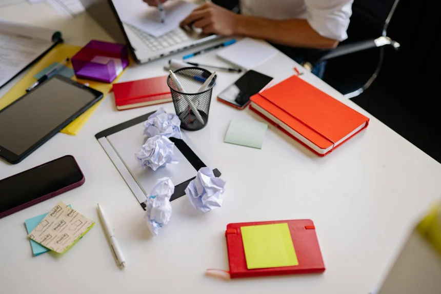 A cluttered desk with crumpled paper, a tablet, colorful notebooks.